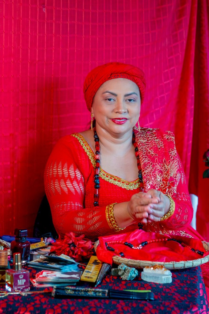 Lisandra de Iansã à mesa com vestido vermelho e dourado, cartas de tarô, cristais e objetos rituais para consulta de tarô amoroso